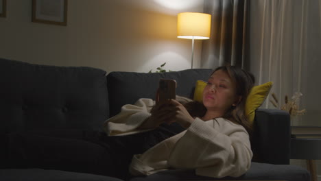 Woman-Spending-Evening-At-Home-Lying-On-Sofa-With-Mobile-Phone-Scrolling-Through-Internet-Or-Social-Media
